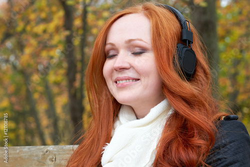 girl portrait, listen music on audio player with headphones, sit on bench in city park, autumn season, yellow trees and fallen leaves