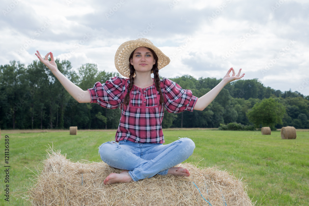 young woman sitting on hay bale meditating in front of a beautiful landscape with blue sky