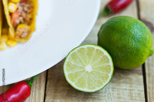 Green lime and white plate with taco, red chili pepper on wooden table. Food ingredient