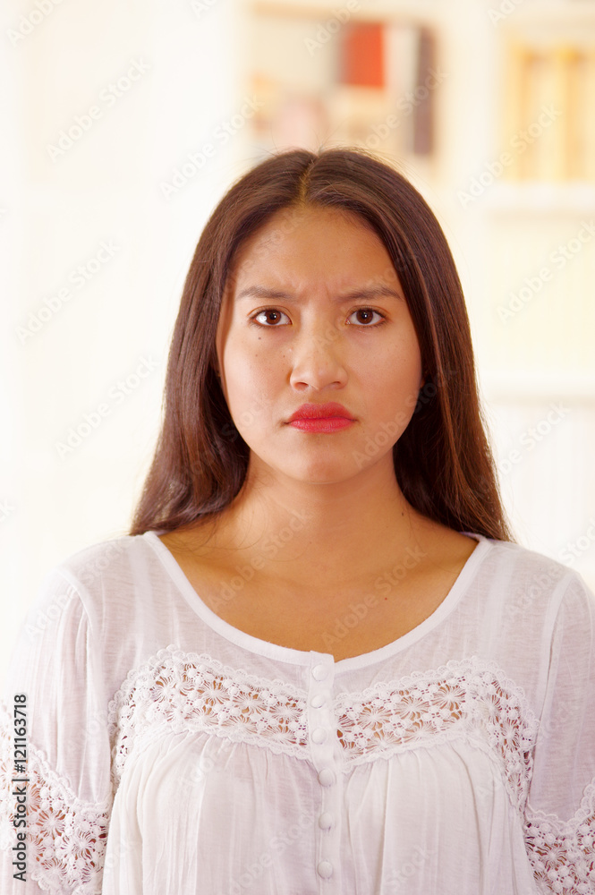 Beautiful young brunette woman wearing white blouse top, facing camera with serious facial expression, bright household background Beautiful young brunette woman wearing white blouse top, facing camera with serious facial expression, bright household background