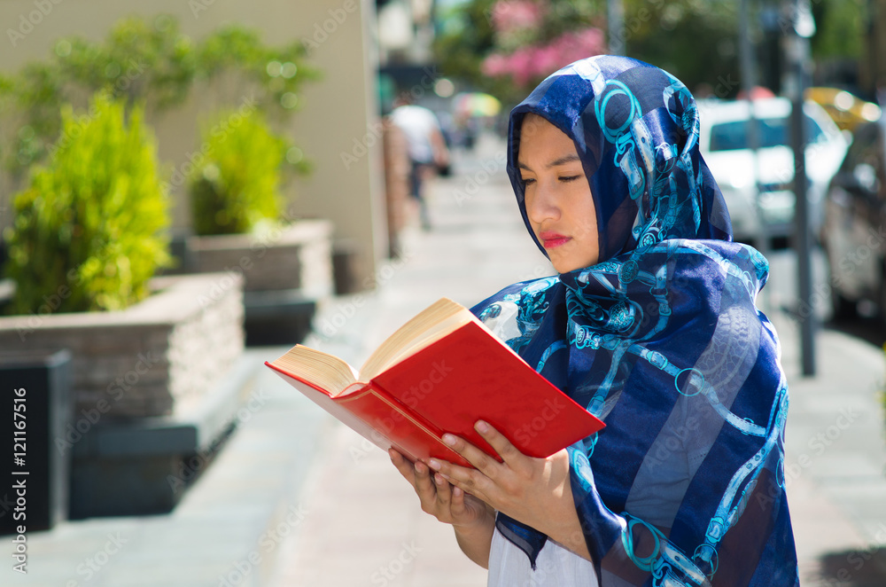 Beautiful young muslim woman wearing blue colored hijab, holding thick reed book and reading in street, outdoors urban background Beautiful young muslim woman wearing blue colored hijab, holding thick reed book and reading in street, outdoors urban background