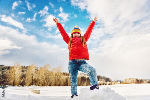 Little girl wearing red jacket and colorful hat, playing with snow in winter time, arms wide open