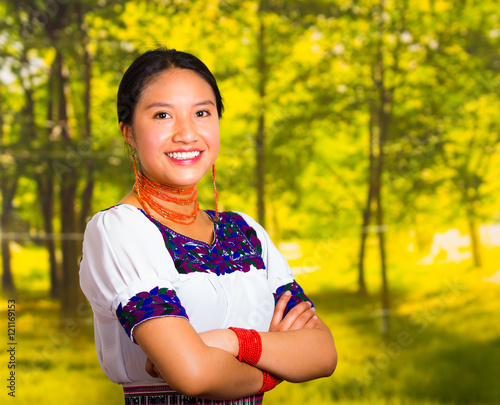 Beautiful young woman wearing traditional andean blouse with necklace, standing posing for camera, arms crossed smiling happily, green forest background