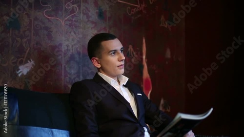 Young businessman in the dark suit and white shirt sitting in the rich interior of the room.