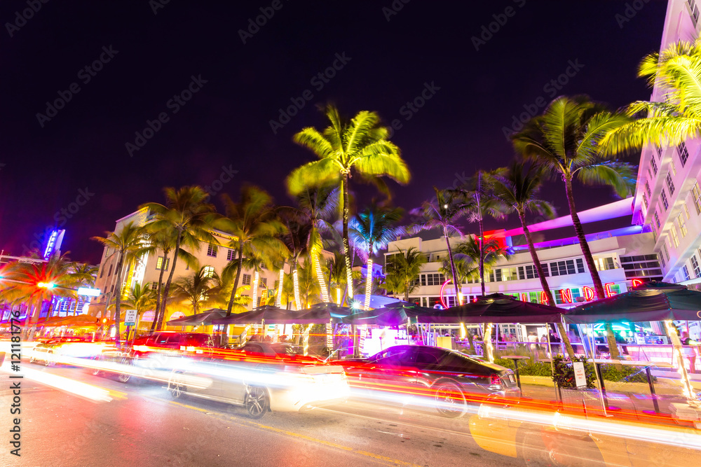 Fototapeta premium Nocna scena Ocean Drive z neonami, palmami, samochodami i dobrze się bawiącymi ludźmi, Miami Beach.