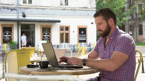 Young man writing on laptop. He is sitting at the table in cafe. He is young and has beard. Man is dressed in checkered shirt. Slider shot right

