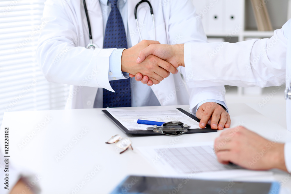 Two doctors shaking hands to each other sitting at the table in hospital office Two doctors shaking hands to each other sitting at the table in hospital office