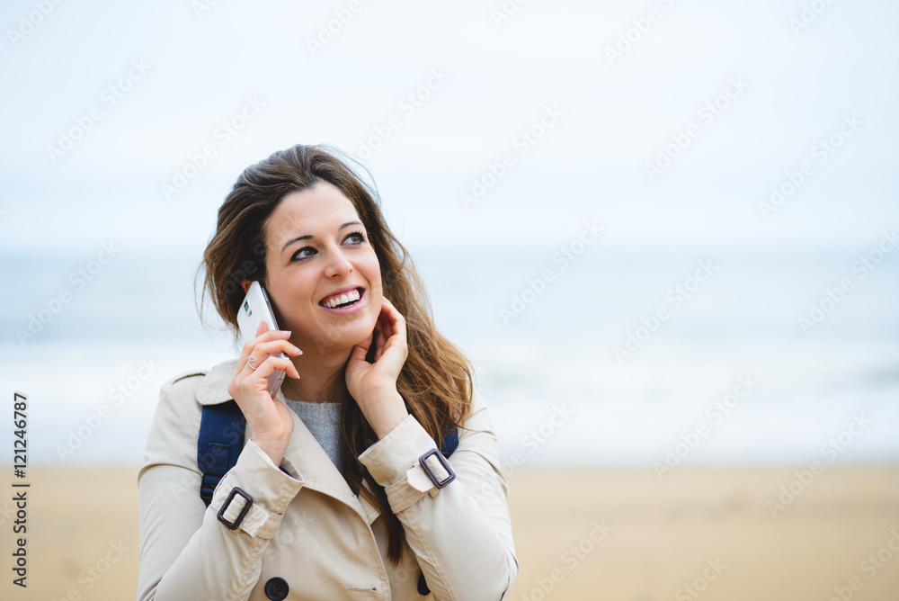 Cheerful woman making a cellphone call during autumn or winter trip to the beach. Female in raincoat using smartphone against the sea. Cheerful woman making a cellphone call during autumn or winter trip to the beach. Female in raincoat using smartphone against the sea.