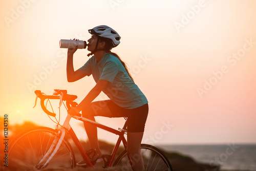 Fototapeta Naklejka Na Ścianę i Meble - Silhouette of female cyclist riding bike and dringking water with sea on background