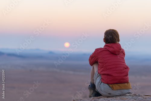Fototapeta Naklejka Na Ścianę i Meble -  Relaxed tourist sitting on rocks and looking at the sunset in the Namib desert, best travel destination in Namibia, Africa. Concept of adventure and traveling people. Rear view, selective focus.