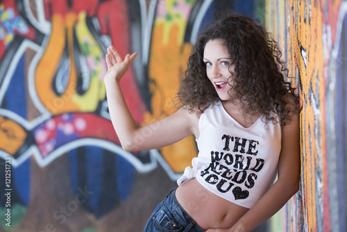 Pretty lady with curly hair wear a crop top shirt and shorts leaning on a graffiti wall