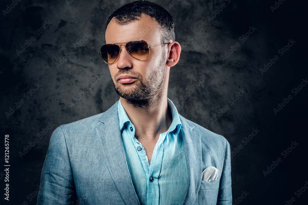 A man in azure jacket and sunglasses posing in a studio.