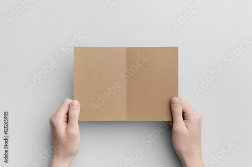 Kraft A6 Bi-Fold / Half-Fold Brochure Mock-Up - Male hands holding a kraft bi-fold on a gray background. © Shablon