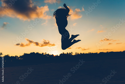 Beautiful silhouette portrait of summer girl jumping on white sand in exotic island at sunset. Serenity, relaxation, mindfulness, stress and carefree concepts.