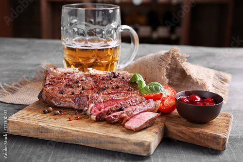 Fotografie Grilled steak with tomatoes on cutting board and mug of beer on wooden table