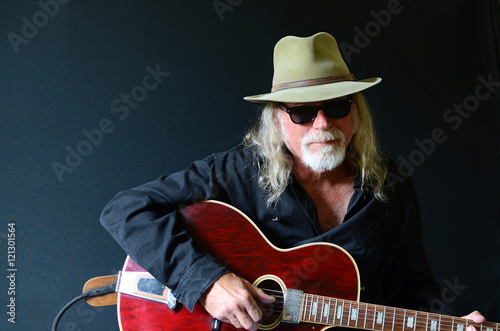 Portrait of a middle aged Caucasian man with long hair and a white beard wearing sunglasses, a fedora hat and a black shirt while holding an electric guitar. Dark background and sidelighting.