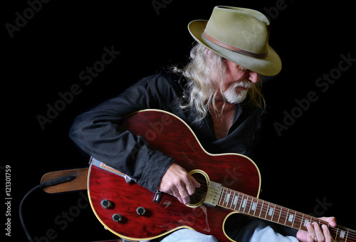 Portrait of a middle aged Caucasian man with long hair and a white beard wearing  a fedora hat and a black shirt while playing an electric guitar. Black background and sidelighting.