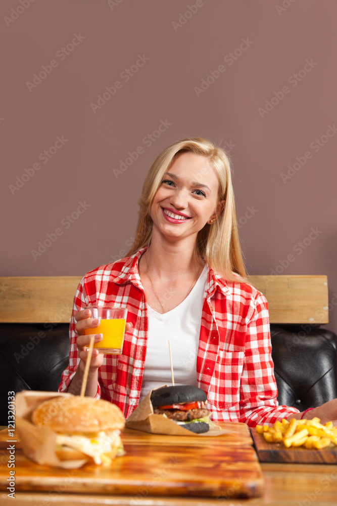 Beautiful Blonde Woman Sitting In Cafe Hold Orange Juice Order Beef Burger Served In Paper On Wooden Table Beautiful Blonde Woman Sitting In Cafe Hold Orange Juice Order Beef Burger Served In Paper On Wooden Table