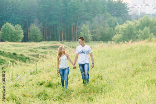A lovely couple walking in a forest area, green grass all around them, holding hands,looking smiling at each other