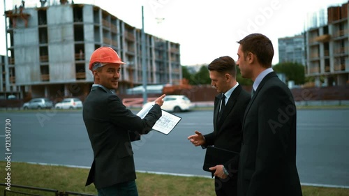 4k. UHD. Three attractive business people, 2 customers andarchitect in helmets with tabletPC and textpad calculation plan discussing about new constructed building at the background with crane and photo