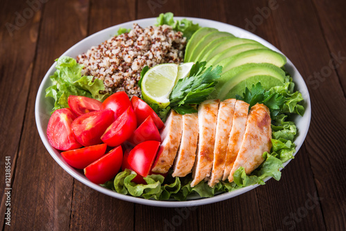 Healthy salad bowl with quinoa, tomatoes, chicken, avocado, lime and mixed greens (lettuce, parsley) on wooden background close up. Food and health.
