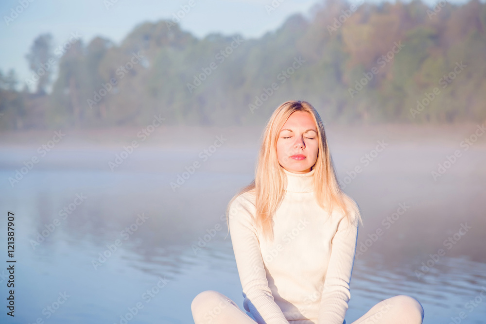 Young, beautiful, gentle woman relaxing and enjoying a first sun rays in the morning in the peaceful place at the lake. Woman practicing yoga early morning. Above the lake is a mist.