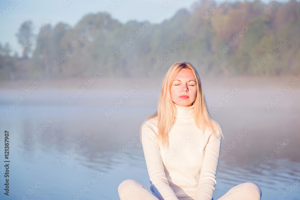 Young, beautiful, gentle woman relaxing and enjoying a first sun rays in the morning in the peaceful place at the lake. Woman practicing yoga early morning. Above the lake is a mist. Young, beautiful, gentle woman relaxing and enjoying a first sun rays in the morning in the peaceful place at the lake. Woman practicing yoga early morning. Above the lake is a mist.