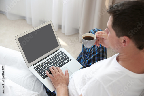 Handsome man sitting on bed with laptop and cup of coffee