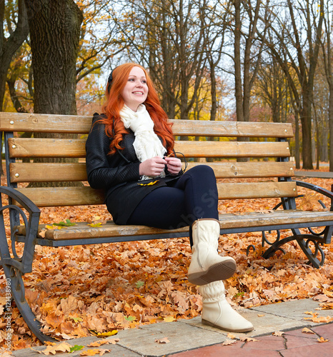 girl in fall season listen music on audio player with headphones, sit on bench in city park, yellow trees and fallen leaves