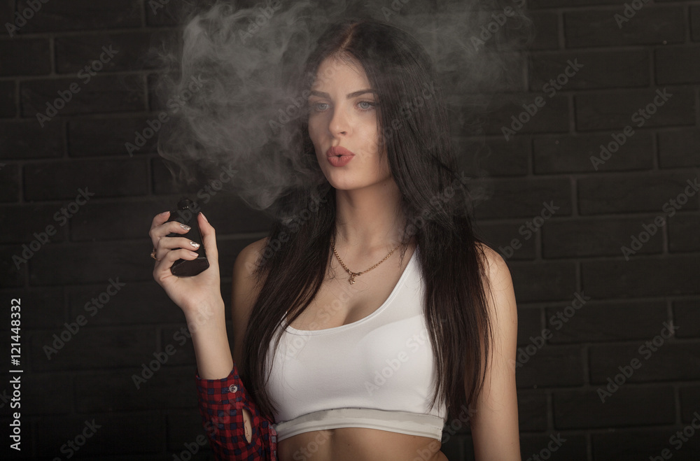 Young beautiful brunette in white jeans shorts woman smoking vaping e cigarette with smoke on black background. Stock Adobe Stock