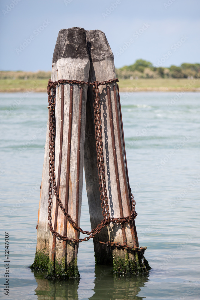 Venecian hisotric poles for mooring ships, sailboats, gondolas. Isolated element. Wooden poles with rusty chains, iron rods. Shore at the background.