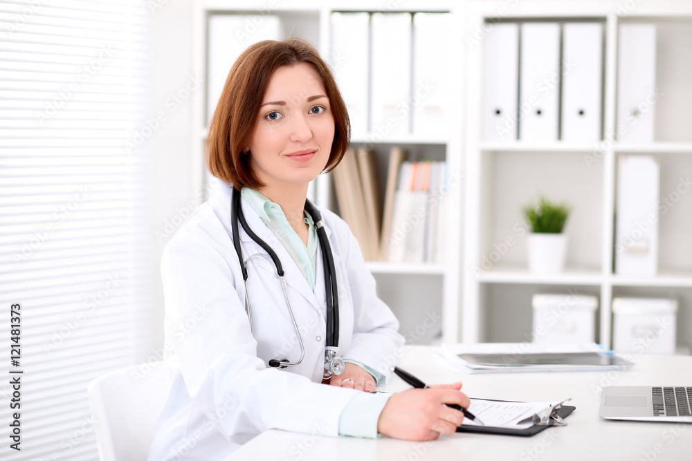 Young brunette female doctor sitting at the table and working at hospital office. Health care, insurance and help concept. Physician ready to examine patient Young brunette female doctor sitting at the table and working at hospital office. Health care, insurance and help concept. Physician ready to examine patient