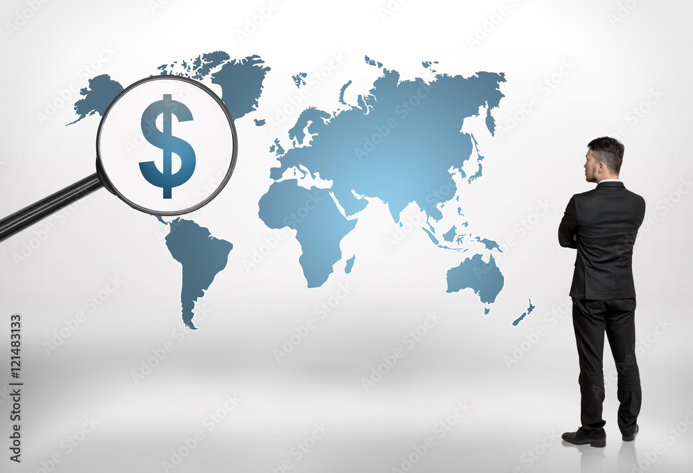 Back view of a businessman looking at world map with big magnifier enlarging dollar sign on it Back view of a businessman looking at world map with big magnifier enlarging dollar sign on it