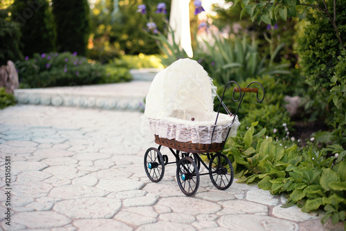 Doll's pram. Vintage doll stroller placed on the stone walkway, alley in a beautiful garden with flowers and trees around. Retro cart dolls made of rattan and white lace