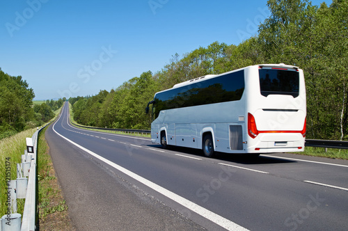 White Bus driving along an empty asphalt road lined with deciduous trees in the countryside