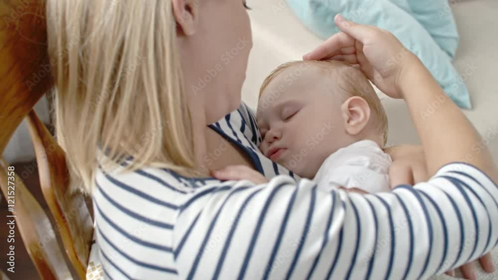 Young woman sitting in rocking chair cradling her cute sleeping toddler and stroking his blond hair Young woman sitting in rocking chair cradling her cute sleeping toddler and stroking his blond hair