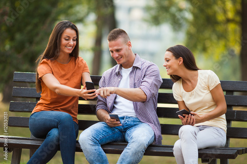 Three friends are sitting on bench in park and using smartphones.
