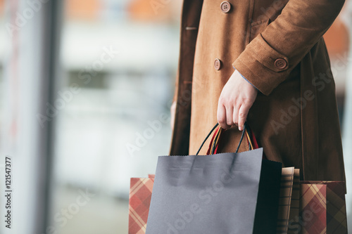 Elegant and modern woman in brown leather jacket or topcoat standing on the city street and holding bunch of shopping bags. Close up shot of woman hand. Lifestyle consumerism theme. 