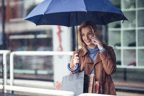 Canvastavla Beautiful, fashionable, young woman standing on the rain with shopping bags and blue umbrella She talking on cell phone and smiling