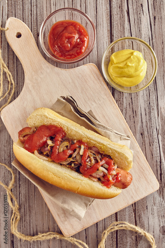Hot dog with onion, ketchup and mustard on a white chopping board on a wooden table. Ketchup and mustard in two transparent bowls.