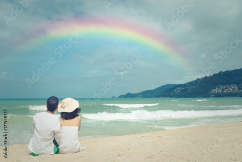 Happy mature couple sitting  on white sand beach enjoying rainbow and sea view