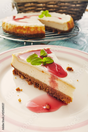 new york cheesecake with cherry sauce on a plate on a blue wooden table, full cake in background photo