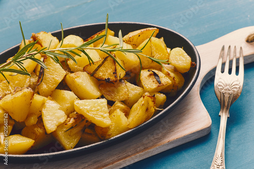 roasted potatoes iwith onion and rosemary in a cast iron pan over a white chopping board on a blue table