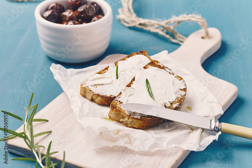 two slice of grilled bread with spreadable cheese and oil on a white chopping board, on a blue wooden table, with olives in a white bowl an rosemary