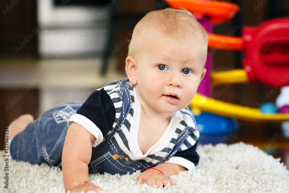 Portrait of cute adorable blond Caucasian smiling baby boy with blue eyes lying on floor in kids children room looking in camera