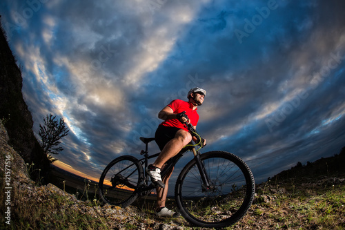 cyclist standing with mountain bike on trail at sunset