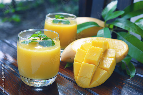fresh mango smoothie with mint leaf on top and ripe yellow mangoes meat in glass cup on wooden background.
