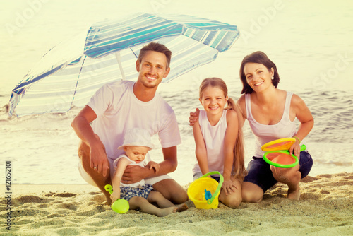 family of four sitting together under beach umbrella on beach
