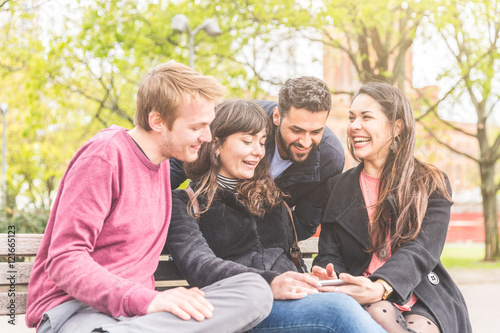 Group of friends having fun at park in Berlin. Mixed race group with caucasian, middle eastern and nordic persons, sitting on a bench and looking at a smart phone. Happiness and friendship concepts.