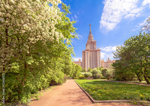 Wide angle view of sunny campus of Moscow university with yellow dandelions and white blossoming apple trees under blue cloudy sky